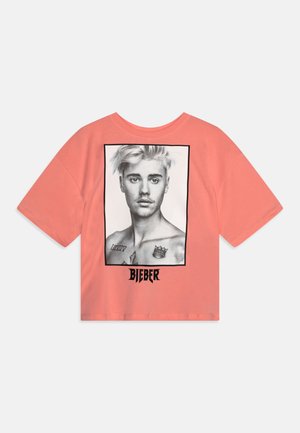T-shirt corail à manches courtes présentant un portrait graphique en noir et blanc au dos, encadré de noir, avec le texte "BIEBER" en dessous.