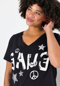 Camiseta negra con el texto blanco "GANG" en estilo desgastado, acentos de estrellas plateadas y símbolos de paz. Tejido suave y casual. Diseño de escote en V.