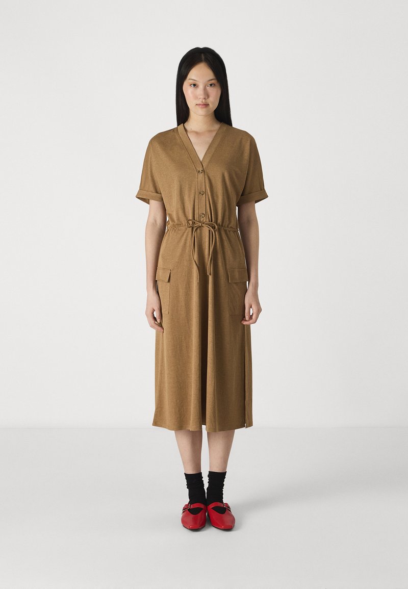 Rochie maro până la genunchi cu mâneci scurte, decolteu în V și închidere cu nasturi. Are două buzunare frontale și un cordon la talie. Purtați cu pantofi roșii și șosete negre.