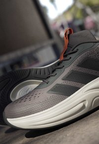 Grauer und schwarzer Laufschuh mit orangefarbenem Zuglasche, weißer dicker Sohle und strukturiertem Obermaterial, schräg gestellt, um die Seiten- und Bodensohle zu zeigen.