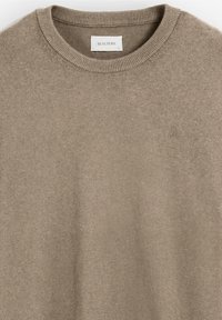 Suéter de punto beige con cuello redondo, collar acanalado y un pequeño logo bordado en el pecho. Textura suave y diseño minimalista.