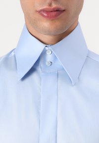 Cămășă din bumbac de culoare albastru deschis, cu un guler structurat, având două nasturi la gât și o textură netedă, evidențiind detalii clare ale cusăturii.