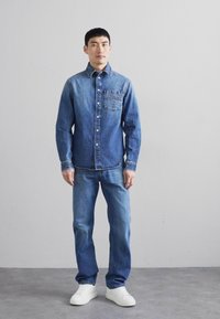 Camicia di jeans con un gradiente che va dal blu chiaro al blu scuro, abbottonata e con una tasca frontale. Indossata con jeans abbinati e sneaker bianche.