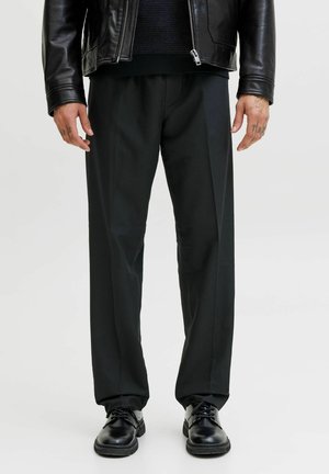 Mann trägt eine schwarze Lederjacke, schwarze Hose und polierte schwarze Schnürschuhe, steht vor einem schlichten hellen Hintergrund.