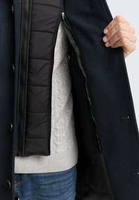Abrigo de color azul oscuro con patrón de espiga y forro negro, que presenta una capa interior con cremallera y un suéter de punto texturizado gris claro debajo.
