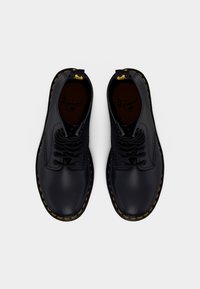 Sorte lædersko med runde tæer, gule syninger og snørebånd samt en indersål med mærke, set oppefra.