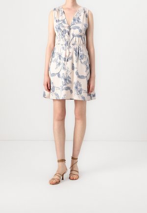 Kvinde iført en ærmeløs, cremefarvet minikjole med blå blomsterprint og beige, remmede hæle, stående foran en ensartet hvid baggrund.