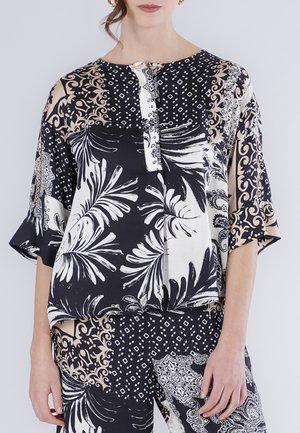 Nainen, jolla on yllään löysä musta, valkoinen ja beige kuvioitu paita sekä siihen sopivat housut, joissa on kukka- ja geometrisia kuoseja.