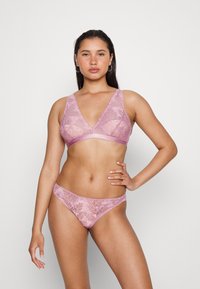 Conjunto de lingerie em renda numa suave cor rosa, apresentando um sutiã com decote em V profundo e uma tanga a condizer, ambos com padrões florais e bordas onduladas.