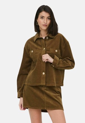 Femme aux cheveux raides et foncés portant une veste en velours côtelé marron avec des boutons dorés et une jupe assortie sur un fond blanc uni.