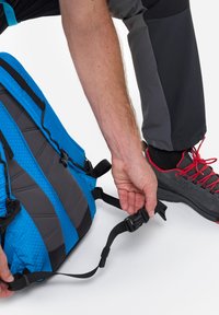 Blauer und schwarzer Rucksack mit strukturierten Paneelen und verstellbaren Trägern, der gerade gesichert wird. Graue Outdoor-Hosen und rot-laced Schuhe sind im Blick.