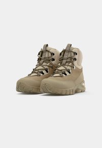 Scarpe da trekking beige con una combinazione di camoscio e tessuto, supporto per la caviglia imbottito, suola in gomma texturizzata e lacci neri a contrasto.