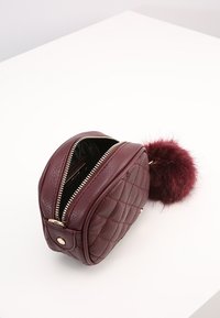 Burgundy leather pouch with quilted pattern, zip closure, and a fluffy burgundy pom-pom accent. Interior is black.