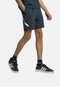 Marineblauwe sportshorts met een witte zijsrand, gecombineerd met zwarte sneakers met drie witte strepen en zwarte sokken. Eenvoudig, functioneel ontwerp.