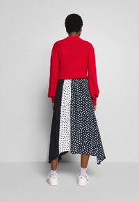 Suéter rojo de punto, mangas largas, patrón abierto en las mangas. La falda tiene paneles en blanco y negro con lunares azules. Zapatillas blancas.