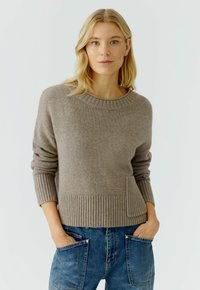 Bruine gebreide trui met een geribbelde halslijn en manchetten, met een voorzak en een relaxed fit, gecombineerd met blauwe denimsalopettes.