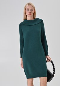 Vestito lavorato a maglia color turchese con un ampio colletto a bavero, maniche lunghe, orlo a coste e una consistenza morbida. Il modello tiene in mano una borsa nera con un manico attorcigliato.