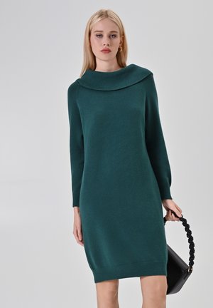 Vestito lavorato a maglia color turchese con un ampio colletto a bavero, maniche lunghe, orlo a coste e una consistenza morbida. Il modello tiene in mano una borsa nera con un manico attorcigliato.