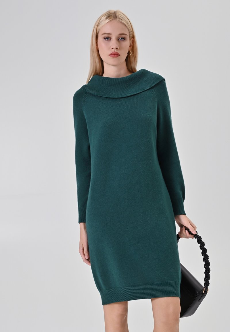 Vestito lavorato a maglia color turchese con un ampio colletto a bavero, maniche lunghe, orlo a coste e una consistenza morbida. Il modello tiene in mano una borsa nera con un manico attorcigliato.