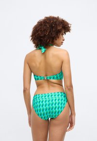 Zaļš modeļa bikini komplekts ar halter tipa topu ar sasienamu lentu, smalkiem metāla akcentiem un atbilstošām zemu jostasvietu biksēm ar ģeometriskiem motīviem.