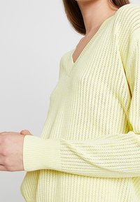 Sweter z dekoltem w serek i długim rękawem w jasnym żółtym kolorze, z delikatnymi pionowymi paskami i otwartą dzianiną, z prążkowanymi mankietami i dolnym brzegiem.