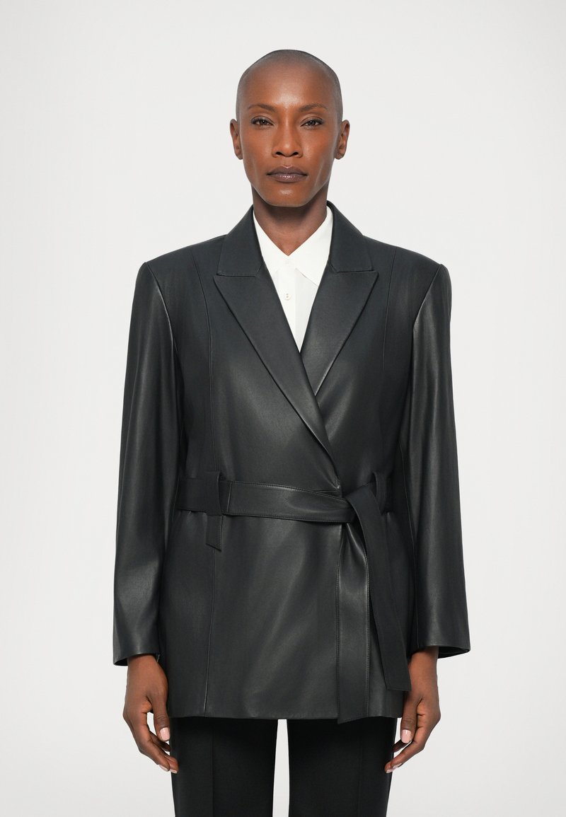 Čierny kožený blazer s prekrývacím dizajnom, dlhými rukávmi a viazaným opaskom. Obsahuje klopový golier a hladkú textúru. Pod ním je biela košeľa s golierom.