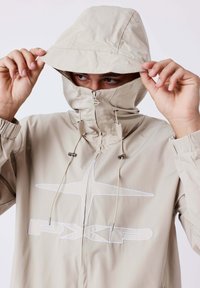 Uomo che indossa una giacca beige con cappuccio e stringhe, parzialmente zipata per coprire il viso, tiene i bordi del cappuccio, logo bianco ricamato sul petto.
