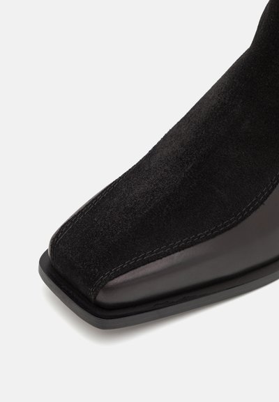 Bottine en suede noir avec un embout carré, des accents en cuir lisse, et des coutures contrastées sur les bords de la chaussure.