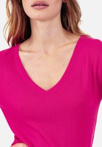 Fuchsia gestrickter Pullover mit V-Ausschnitt, der eine weiche Textur und eine eng anliegende Silhouette aufweist. Sichtbare Details an Schultern und Ausschnitt.