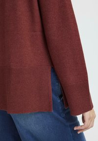 Un pull en tricot couleur rouille avec un ourlet côtelé et des fentes latérales, doté de longues manches et d'une coupe décontractée, associé à un jean en denim.