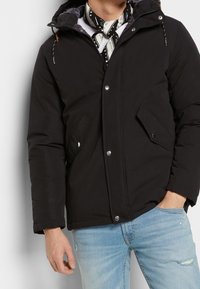 Svart puffjacka med huva, tryckknappar och två framfickor. Texturerat tyg med kontrasterande foder och dragskoaccenter.