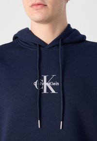 Felpa con cappuccio blu navy, dotata di coulisse e logo "Calvin Klein" ricamato in argento sul petto. Tessuto morbido e vestibilità regolare.