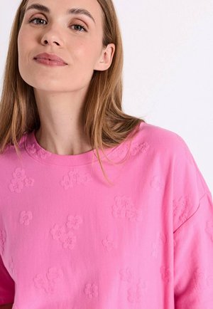 Jonge vrouw met lichtbruin haar, gekleed in een felroze sweatshirt met gestructureerde bloemmotieven op een witte achtergrond.