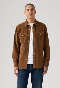 Bruine ribfluwelen shirt met twee borstzakken en drukknoopsluiting, gedragen over een wit T-shirt, gecombineerd met donkerblauwe jeans.