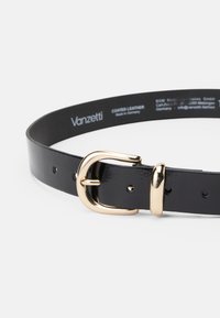 Curea din piele vopsită în negru, cu o cataramă aurie, având o finisare netedă și mai multe laturi pentru ajustarea dimensiunii. Logo-ul este imprimat pe partea interioară.