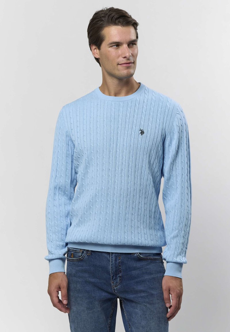 Lyseblå strikket genser med kabelmønster, ribbestrikket rund hals og mansjetter. Liten mørk emblem på brystet. Brukt med blå jeans.