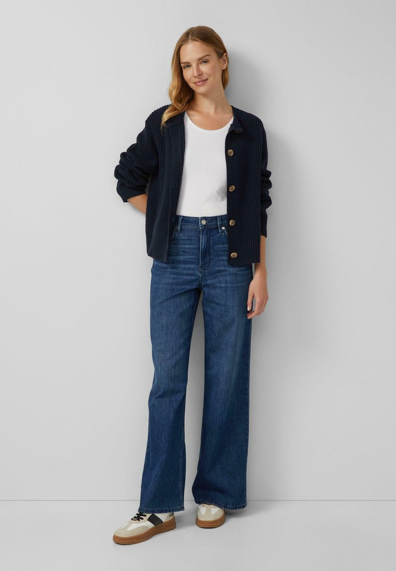 Marineblauwe geribbelde cardigan met knopen, witte crewneck top, hoge taille brede spijkerbroek van donkerdenim en beige sneakers met zwarte accenten.