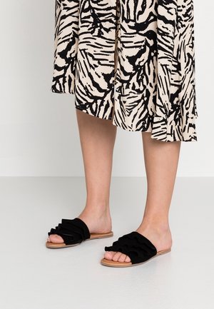 Sandales noires à glissière avec une tige en tissu doux ornée de volants. Semelle plate de couleur naturelle, avec une texture lisse.