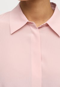 Helle rosa Bluse mit glatter Oberfläche, spitzem Kragen und einer Knopfleiste vorne, die subtile Steppdetails auf dem Stoff aufweist.