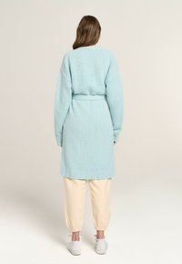 Light blue, knitted cardigan with long sleeves, fitted belt at the waist, and ribbed hem. Paired with cream tapered pants and white sneakers.