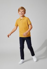 Żółta koszulka polo z zapięciem pod szyją, w połączeniu z ciemnoniebieskimi spodniami i białymi trampkami. Ubrania mają luźny krój i gładką teksturę.