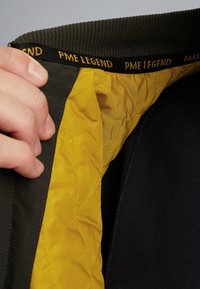 Intérieur de veste vert foncé avec doublure matelassée jaune et accents noirs. Le col présente une bande de marque avec un texte doré.