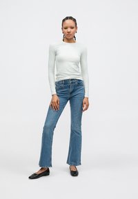 Ανοιχτό γκρι μακρυμάνικη μπλούζα, καμπανούλες μπλε τζιν και μαύρα επίπεδα παπούτσια. Η μπλούζα έχει εφαρμοστή σχεδίαση· το τζιν διαθέτει κλασική υφή denim και ραφές.