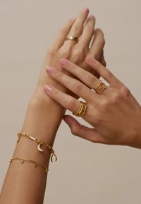 Joyas de oro en las manos, que incluyen múltiples anillos, una pulsera texturizada con un charm en forma de media luna, y otra pulsera con pequeños acentos circulares.