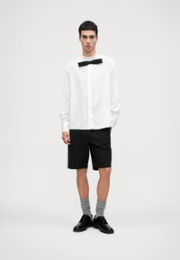 Bílá košile s volánkovým límečkem a černým motýlkem, v kombinaci s černými šortkami, šedými ponožkami a černými botami, na neutrálním pozadí.