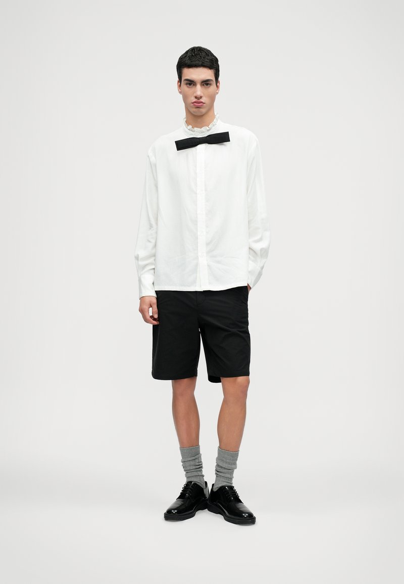 Bílá košile s volánkovým límečkem a černým motýlkem, v kombinaci s černými šortkami, šedými ponožkami a černými botami, na neutrálním pozadí.