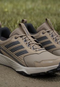 Tan athletic shoes with gray accents, textured upper, lace system, cushioned ankle support, and a rugged sole, labeled "CLIMA PROOF."