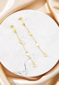 Goldene Tropfenohrringe bestehen aus verknüpften Ketten mit runden Akzenten, von denen einer mit einem Steindetail versehen ist, und sind vor einer marmorierten, runden Oberfläche angeordnet.