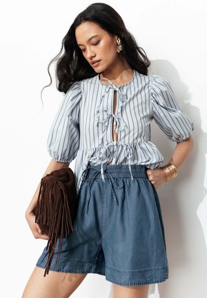 Femme portant un chemisier rayé à manches bouffantes avec liens à l’avant, short en jean taille haute, grandes boucles d’oreilles dorées, bracelet manchette, tenant un sac à franges marron.