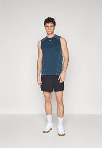 Blå ermeløs trenings-topp med lysegrå detaljer, kombinert med svarte shorts. Modellen har på seg hvite joggesko og hvite sokker til leggen.